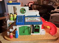 Front and side view of the Sesame Street Clubhouse playset showing blue roof, green doors, red slide, and yellow broken swing on tan base with sticker details intact.