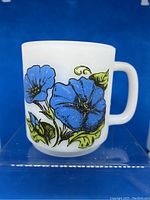 Photo showing side of milk glass mug with bright blue morning glory flowers and green leaves against white background.