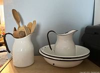 Two white enameled jugs and two bowls displayed on shelf, one jug holds wooden utensils
