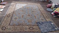 Full view of large area rug with light blue center, beige and brown floral patterns and ornate border. Smaller welcome mat is placed on top showing contrast in design and color.