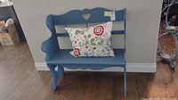 Blue painted wooden bench with heart cutout backrest, curved armrests, and scalloped edges shown with an embroidered floral throw pillow.