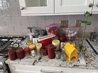 Wide view of 24+ pieces including various candles in red, cream and green colors, candle holders made of ceramic, glass, and metal, plus miscellaneous coasters and a unique yellow house-shaped candle holder.