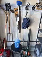Photo shows multiple garden tools including pitch fork, rake, broom, hand saw, umbrella and leaf blower, and a blue plastic fluid container.