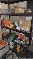 Shelving unit with various gardening and hardware supplies including baskets, boxes, fencing rolls, hose, motor oil container, and cleaning sprays.
