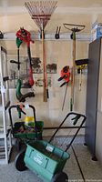 Photo showing two handheld weed eaters hung on wall, two seed spreaders on floor, and various garden hand tools hung on wall including rakes and hoe.