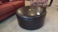 Round dark brown leather storage ottoman closed top, showing four stitched sections.