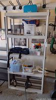 Front view of white plastic five shelf storage unit with various boxes and items on the shelves, background includes garage wall and hanging tools.