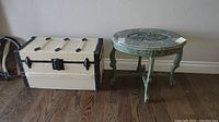 Overall view of painted cream trunk next to green carved side table on wooden floor