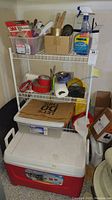 Image showing a white wire storage shelf on wheels with various hand tools, duct tape, spray bottle of Rain-X glass cleaner, disposable gloves, paper towels, and plastic bin with lid on top of a red and white Igloo cooler.