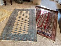 Two Asian-inspired wool rugs lying on carpeted floor. One cream colored with blue/red patterns, the other dominantly red patterned.