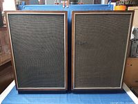 Pair of large rectangular speakers with dark fabric grilles framed with wood trim on front, standing side by side.