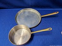 Two copper pans viewed top side showing stainless steel interiors and brass handles on a blue background.