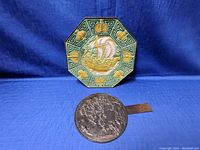 Two Asian metal dishes on blue fabric background; an octagonal colorful embossed dish and a round dark embossed dish with handle.