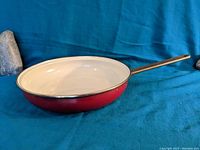 Side view of cherry red enamel frying pan with cream enamel interior and long brass handle.