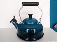 Blue enamel Le Creuset tea kettle front view showing round body, spout with black lever and black handle.