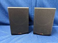 Pair of Polk Audio R-150 speakers with black fabric front grilles on, positioned side by side against blue fabric background.