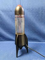 Full view of the rocket-shaped lamp showing black base and top, clear center with water and glitter, cord attached