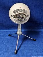 Blue Snowball ICE USB microphone on an adjustable silver tripod stand against a blue background. Shows front angle with Blue logo and red indicator light visible.