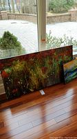 Two paintings leaning against a window with hardwood floor. Larger 25x48 inch abstract floral artwork visible in red, green and yellow hues. Smaller 15x18 inch signed landscape painting partially visible, showing nature scene with sunset and trees.