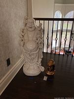 View of the large white ceramic Buddha sculpture with small children figures around it, and the smaller bronze Pier 1 Buddha sculpture beside it on a dark wooden floor.
