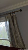 Photo of light-colored drapes with horizontal stripes hung on an ornate curtain rod inside a room, showing part of the window and wall.