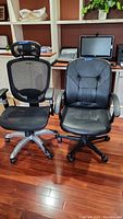 Two black office chairs side by side on wood floor in office setting. One chair is mesh with headrest, the other faux leather with visible wear.