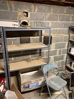 Photo of one metal shelving unit with dirt and dust on wooden shelves, located against basement cinder block wall with debris below.