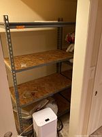 First metal shelving unit showing three wood particle board shelves with some debris and a small white heater/dehumidifier on the floor in front.
