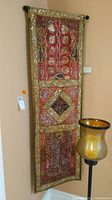 Front view of one ornate sequined fabric wall tapestry showing detailed patterns and sequins, hung on a black rod on a beige wall near a decorative candle holder.