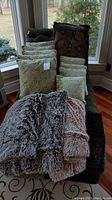 Photo showing four fuzzy throw blankets in varying shades of brown and beige, alongside nine swirl pattern pillows in green tones.