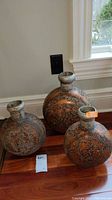 Three round metal vases with floral embossed decoration placed on hardwood floor by a wall outlet.