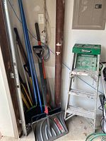 Photo showing collection of yard tools including snow shovels, dirt shovels, and a coiled pool cleaner hose against a garage wall.