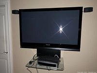 Panasonic 55 inch Viera TV mounted on black stand with glass shelf holding an electronic device beneath the TV.