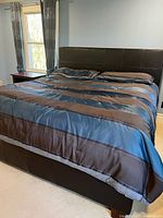 Front angled view of king sized bed with dark brown leather upholstered frame and headboard, mattress made with striped blue and brown bedding, near window with curtains.