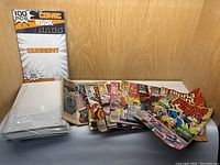 Pack of 100 comic book sleeves in original packaging along with a collection of comic books spread out on surface.