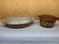 A photo showing a large divided oval casserole dish with a frosted glass lid next to a small round casserole with a brown lid, both featuring golden-yellow Early American patterns on a brown base.