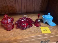 Lot of four Fenton glass pieces displayed on wooden shelf: ruby red covered jar, ruffled bowl with metal handle, hobnail bell, and blue opalescent hobnail shoe.