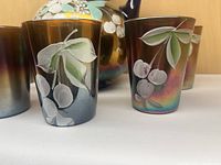 Close-up showing two iridescent glasses decorated with hand painted white flowers and green leaves in front of the pitcher body.