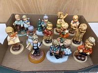 Overall view of 12 Hummel figurines in a cardboard box, showing grouping and details of each figurine.