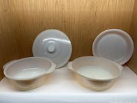 Four vintage Fire King pieces: two round casserole dishes (one with lid), and a pie pan, all white glass, shown on shelf