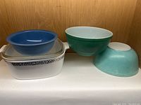 Set of miscellaneous bowls and bakeware including Pyrex blue bowl with lid, white rectangular casserole dish with decorative border, and various ceramic mixing bowls in green and turquoise.