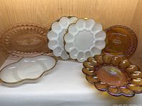 Six pieces of vintage FireKing and pink glass including milk glass egg trays and assorted serving plates with gold trim and starburst patterns, arranged in a wooden shelf.