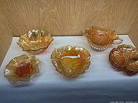 Front view of the seven vintage carnival glass pieces showing their shapes and iridescent amber-orange coloration on a white surface with a wood background.