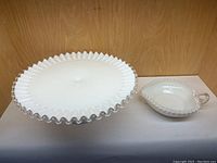 Front view of milk glass cake stand and matching bowl with handle, both with silver lace ruffled edges.