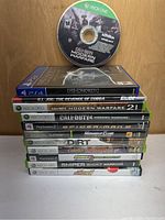 Stack of 12 video game cases with one Xbox One loose disc (Call of Duty Modern Warfare) on top, showing titles and platform details.