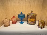 Full display of five vintage glass items on wooden shelf: large hand-painted Fenton jar, three additional lidded jars in amber, orange-red, and blue, and an iridescent small vase.
