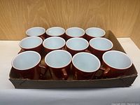 Lot of 12 brown Pyrex mugs placed in a cardboard tray showing white interiors.