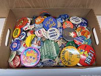 Photo showing a large box filled with assorted vintage buttons, mainly featuring text and images related to political campaigns and safety messages.