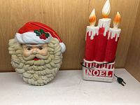 Singing Santa face and light-up candle Noel blow molds shown side by side on wooden surface.