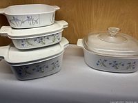 Stacked four white CorningWare casserole dishes with blue and purple floral pattern with three lids, two plastic and one glass.